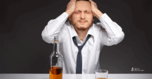 Stressed man in a dress shirt and loosened tie holding his head, sitting in front of a bottle and glass of alcohol, symbolizing the emotional toll of alcohol addiction.