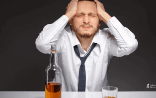 Stressed man in a dress shirt and loosened tie holding his head, sitting in front of a bottle and glass of alcohol, symbolizing the emotional toll of alcohol addiction.