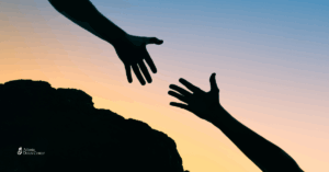 Silhouetted hands reaching out to each other against a sunset, symbolizing support, connection, and offering help to someone in need.