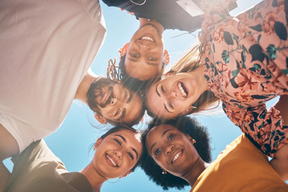 A group of friends forms a tight circle, smiling down at the camera against a bright blue sky.
