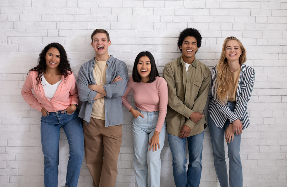 A diverse group of five young adults stands against a white brick wall, smiling and laughing, exuding a sense of friendship and positive energy.