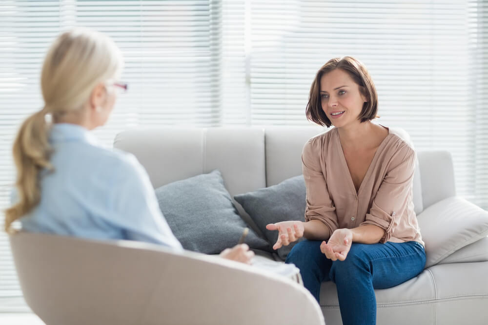Two women are seated on a couch having a relaxed and open conversation, suggesting a counseling or therapy session in a comfortable, private setting.