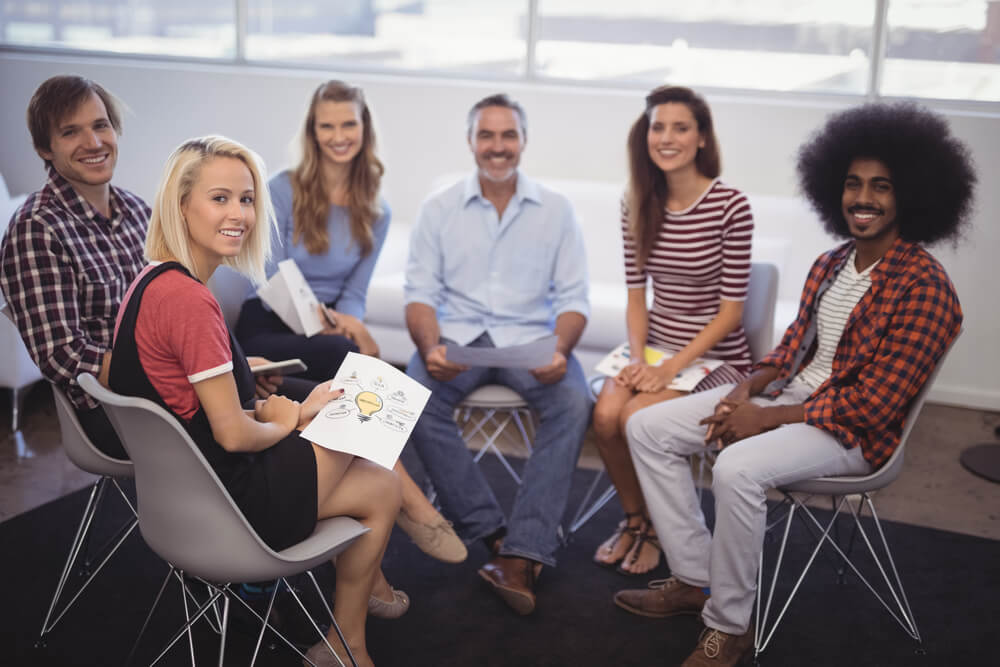 A group of people sit in a circle with papers in their hands while smiling at the camera,