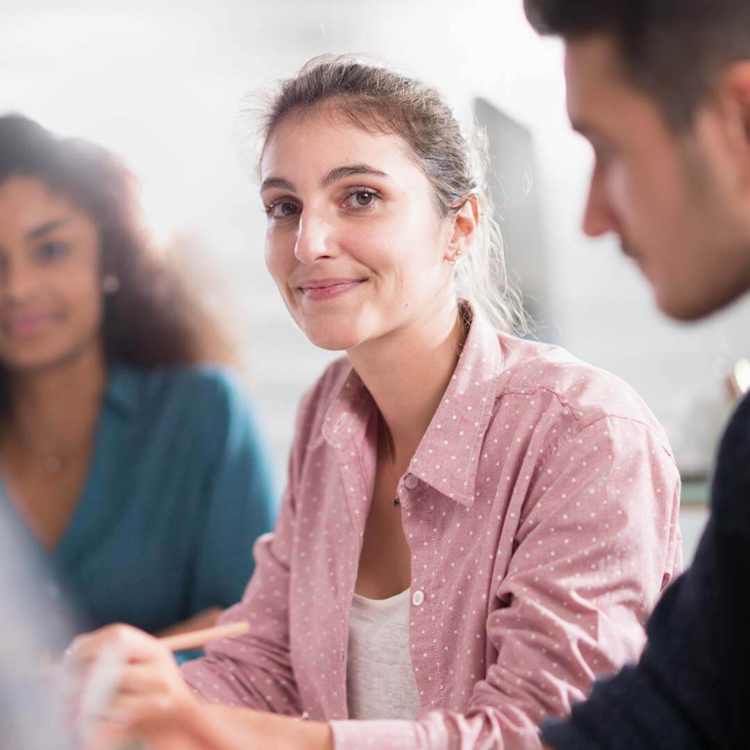 A young woman in a pink shirt smiles softly at the camera while sitting in a group setting, conveying a sense of calm and confidence amidst a collaborative discussion.