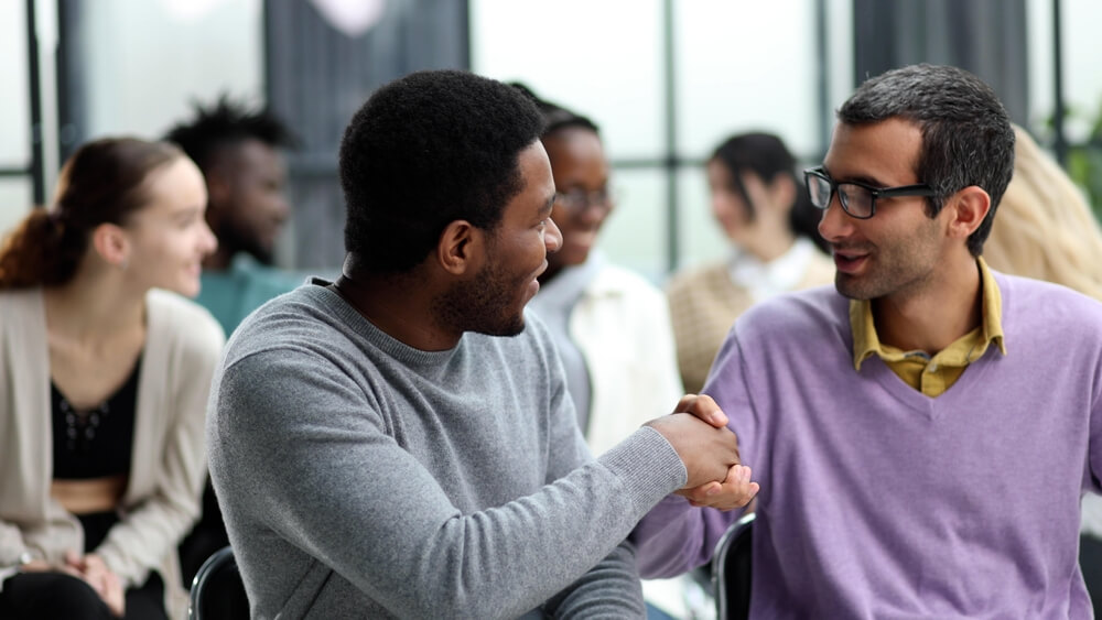 Two men shake hands amid a group of people in the background.