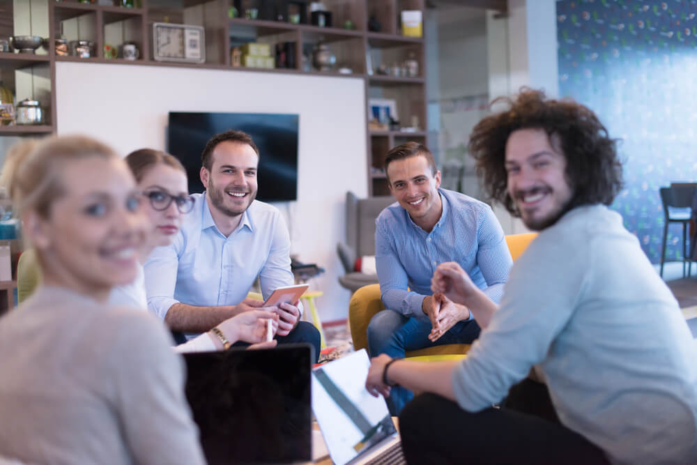 A group of young professionals sits in a casual office setting, smiling and engaged in a discussion, reflecting a collaborative and friendly work environment.
