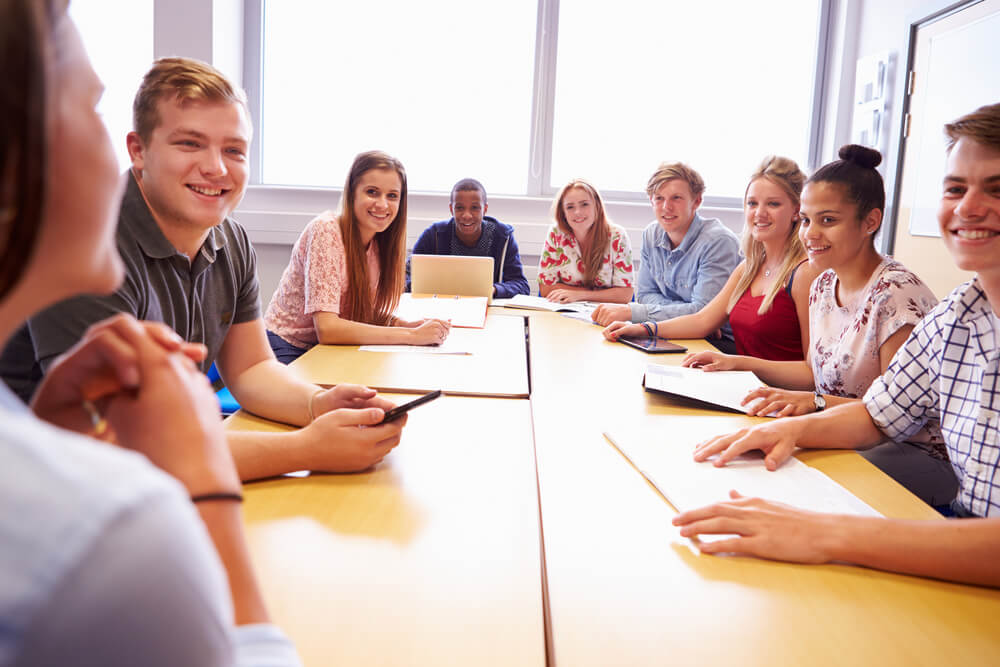 A group of young adults at a conference table stare at a woman at the head facing away from the camera.