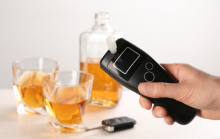 A breathalyzer is held up in front of two glasses and a bottle of alcohol, and a car key.