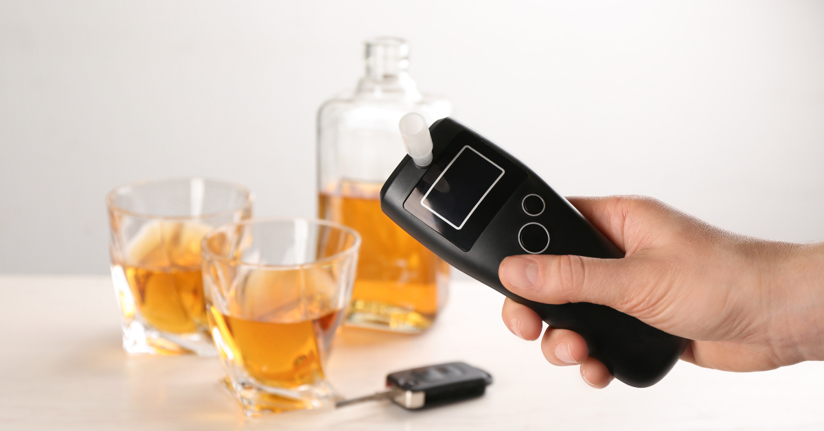 A breathalyzer is held up in front of two glasses and a bottle of alcohol, and a car key.