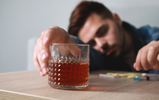 A man sits passed out at a table with various drugs scattered, his hand reaching for a glass of dark liquid.