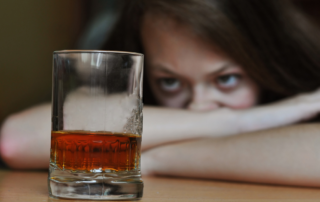 A woman sits at a table with his face in her arms, glaring at a glass of dark liquid.