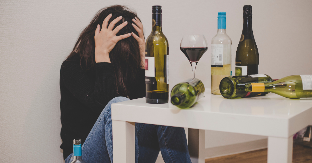 A woman sits covering her head in shame next to a small table with several wine bottles.