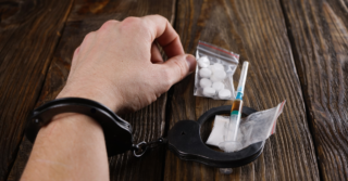 Close-up of a hand in handcuffs next to heroin-related drug paraphernalia, including a syringe and baggies of white powder and pills, symbolizing the link between heroin dependence and legal consequences.