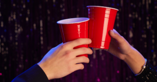 Two hands holding and clinking red plastic cups, set against a dark background with purple lights.