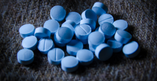A pile of blue pills scattered on a dark surface. The pills are round with a line dividing them in half.