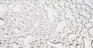 Water droplets on a smooth surface, possibly glass or metal. The droplets vary in size and create an abstract pattern.
