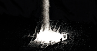 A stream of white powder being poured onto a dark surface. The powder is spreading as it lands, creating a dramatic contrast against the dark background.