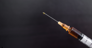 Close-up of a syringe with a needle, filled with a brownish liquid, against a blurred dark background. A drop of liquid is visible at the tip of the needle.