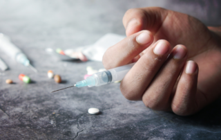 A hand holding a syringe, with scattered pills and another syringe in the background, suggesting drug use.