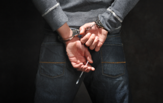 A person in handcuffs is shown from behind, holding a syringe in their hands, suggesting involvement with drug use and law enforcement.