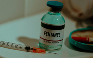 A vial labeled "Fentanyl" for injection alongside a syringe on a white surface, representing medical or illicit use of the drug.