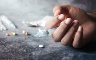 A hand holding a syringe, surrounded by scattered pills and drug paraphernalia on a concrete surface.