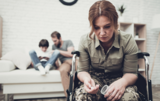 A woman in military uniform sitting in a wheelchair, looking down at a pill bottle, while a man and child sit in the background on a couch.