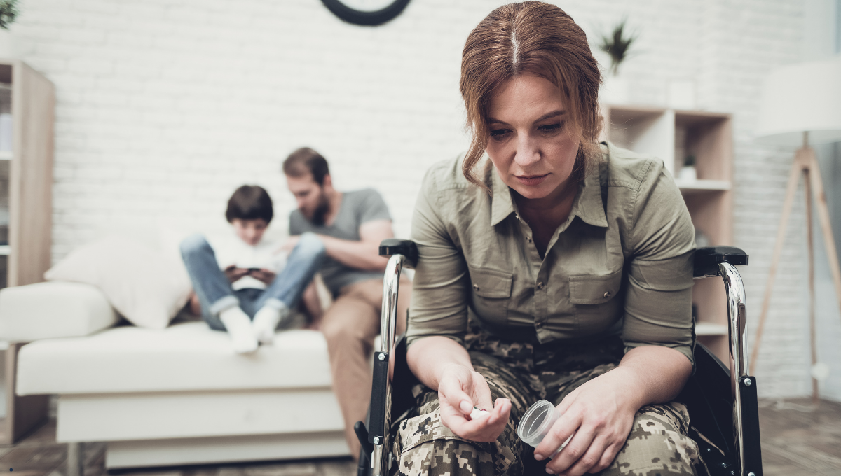 A woman in military uniform sitting in a wheelchair, looking down at a pill bottle, while a man and child sit in the background on a couch.