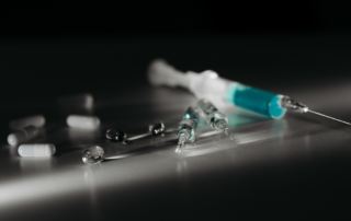 Close-up of medical syringes filled with blue liquid, surrounded by scattered white capsules and clear capsules on a dark surface, under dim lighting.
