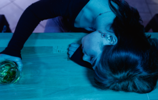 Woman with her head resting on a table holding a glass of alcohol, depicting exhaustion or effects of drinking.