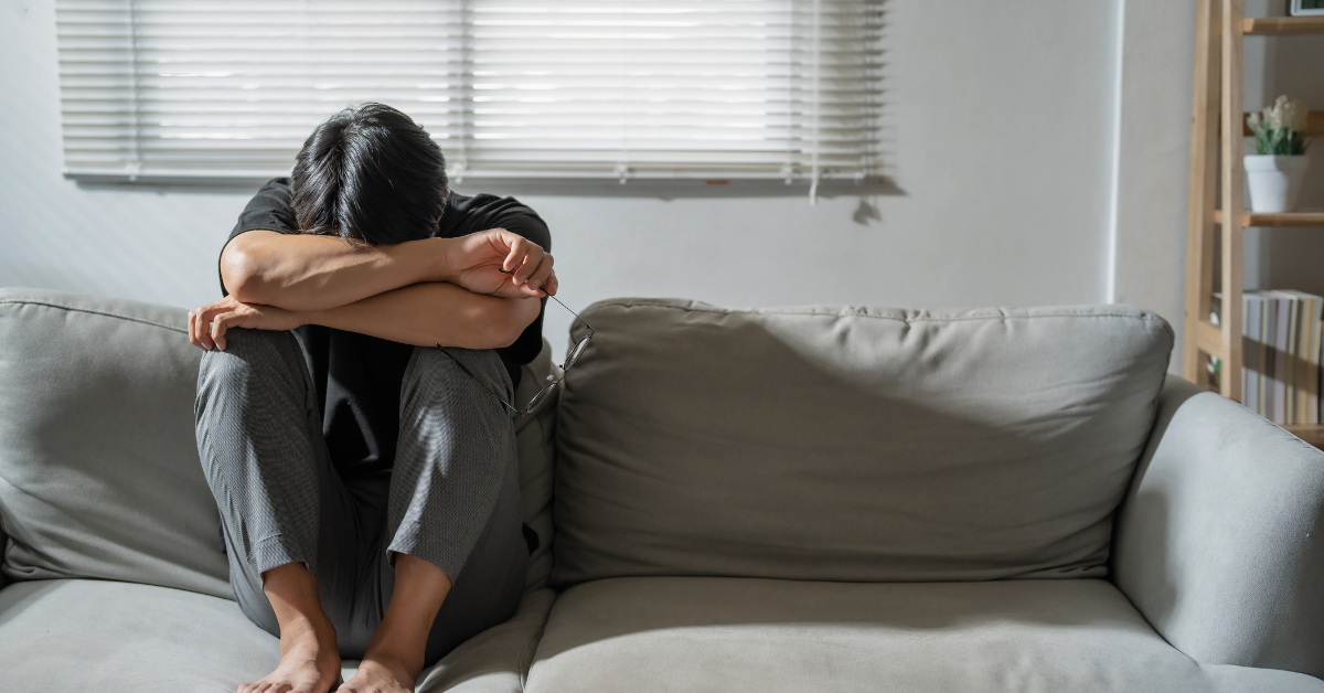 A person sitting on a gray couch with their knees up, arms crossed over their face, appearing upset or reflective in a softly lit room with blinds and bookshelves in the background.