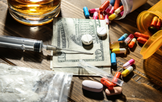 Various drugs, pills, a syringe, and a glass of alcohol placed on a wooden table alongside cash, symbolizing the challenges of addiction.