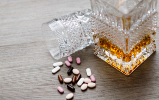 A close-up of a spilled crystal glass next to a bottle of amber liquid, resembling alcohol, with various colorful pills scattered on a wooden surface.