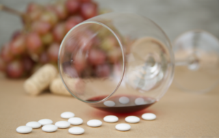 Spilled glass of red wine with white pills scattered on a table, with grapes and corks blurred in the background.