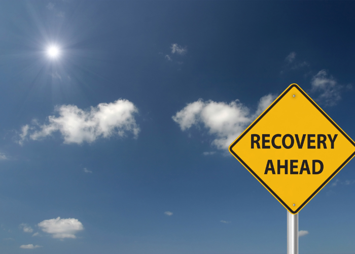 Yellow road sign with the words 'Recovery Ahead' against a bright blue sky with scattered clouds and sunlight.