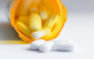 Close-up of an open prescription pill bottle with yellow and white pills spilling out on a white surface.