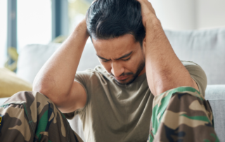 Veteran in camouflage pants sitting on the floor with his head in his hands, showing signs of emotional distress related to PTSD and addiction.