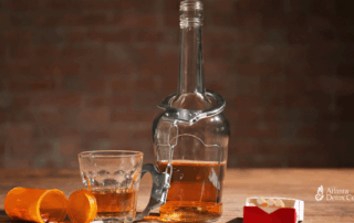 Glass and bottle of liquor next to spilled prescription pills and a cigarette box, symbolizing the dangers of mixing Wellbutrin with alcohol.